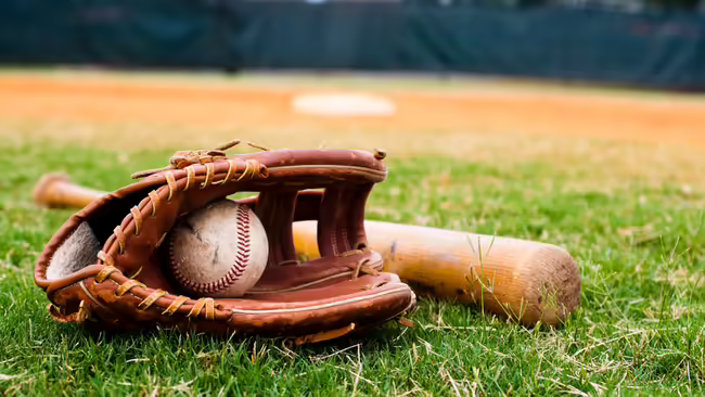 Baseball glove holding a baseball with a wooden bat resting beside it on the grass of a baseball field, with the infield visible in the background.