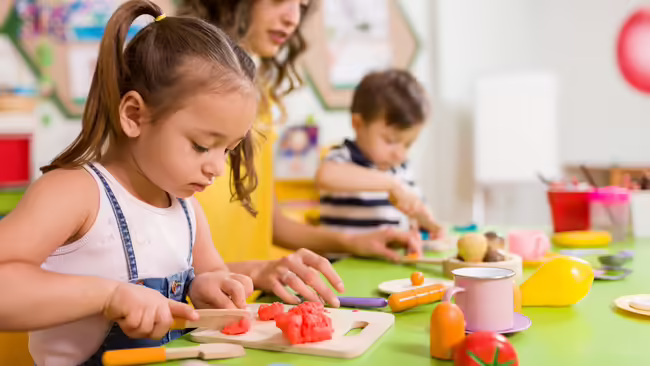Children at a play table using toy food and utensils to pretend-cut vegetables during a kitchen-themed activity.
