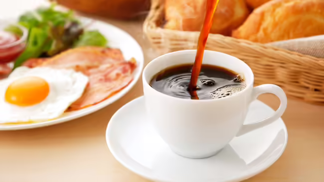 Coffee being poured into a coffee mug with a plate of breakfast food on the table.