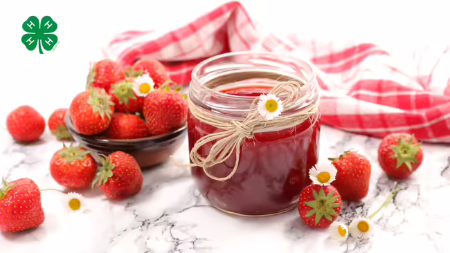 A jar of strawberry jam with strawberries on the table. A green 4-H logo in upper left corner.