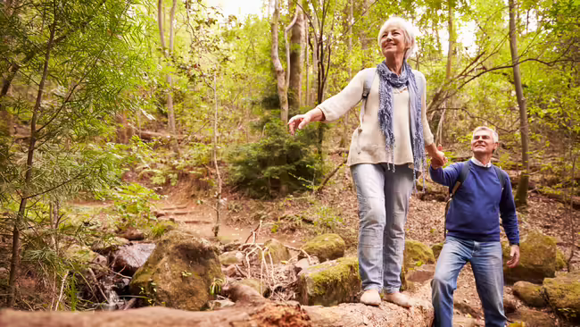 An older woman balancing on a log while a man holds her hand.