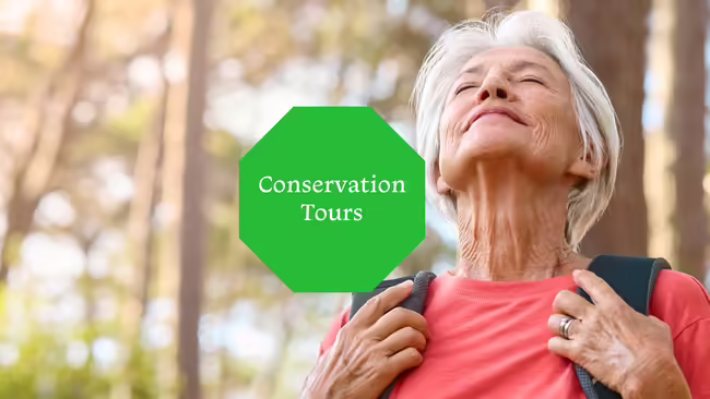 An older woman standing in the woods with her head tilted back and her eyes closed enjoying the sun on her face.