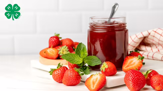 A jar of strawberry jam with strawberries on the table. A green 4-H logo in upper left corner.