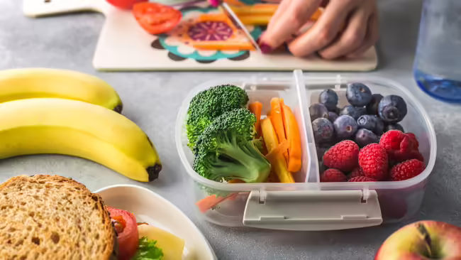 A table with a lunch container, fruit, sandwich, cutting board, and a person cutting a vegetable on the cutting board. Lunch container contains vegetables and fruits.