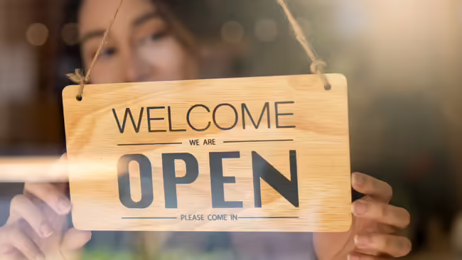A person hanging up an open sign that reads welcome we are open, please come in.