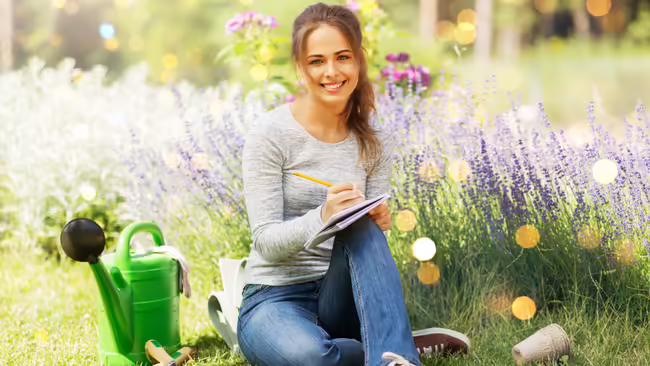 A woman sitting on the grass in a garden, writing in a notebook next to a green watering can and purple flowers.