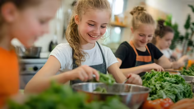 A group of children wearing aprons participate in a cooking class, preparing salads.