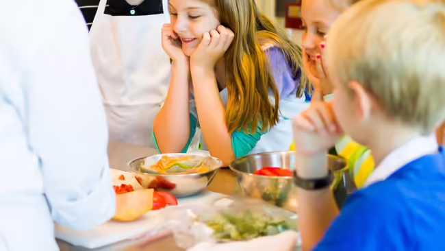Kids wearing aprons watching someone mix ingredients in a bowl.