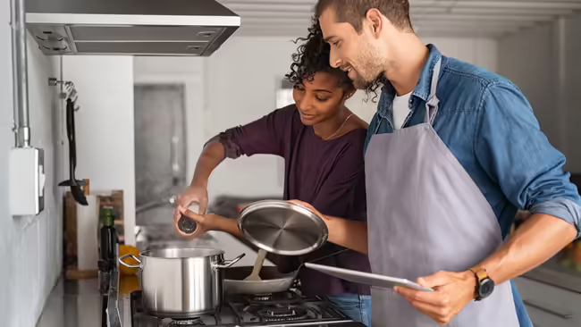 Woman adding salt in pot while cooking while a man looks over her shoulder holding a pan lid.