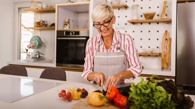 A woman with an apron stands in a bright kitchen, chopping fresh vegetables on a cutting board.