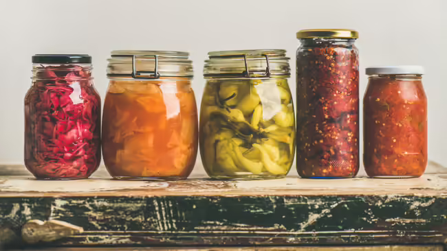 Five glass jars of preserved foods arranged on a rustic wooden table.
