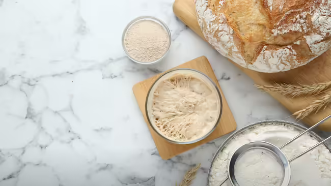 A white marble surface, including a jar of active starter, a bowl of flour with a sifter, a bowl of yeast, wheat stalks, and a freshly baked sourdough loaf.