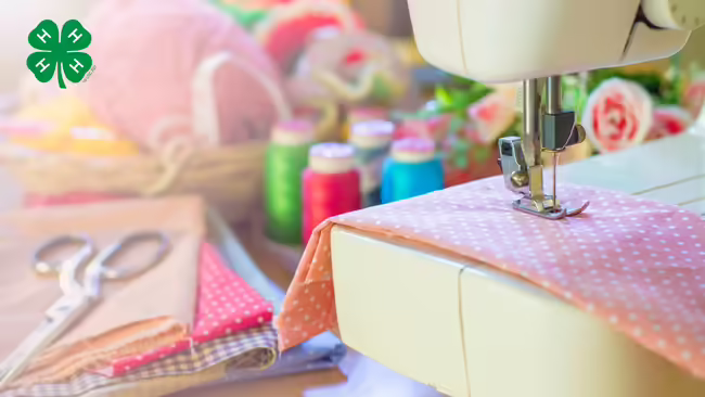 A sewing machine stitching fabric, surrounded by spools of thread, scissors, and fabric swatches, with the green 4-H logo in the upper-left corner.