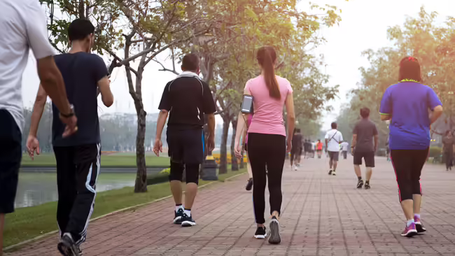 People walking for exercise along a wide, tree-lined park path beside a pond.
