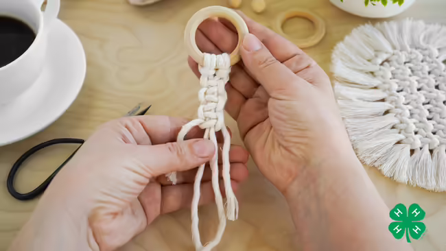 Hands crafting a macramé design attached to a wooden ring on a wooden tabletop next to a coffee cup and scissors. Green 4-H clover in bottom right corner.