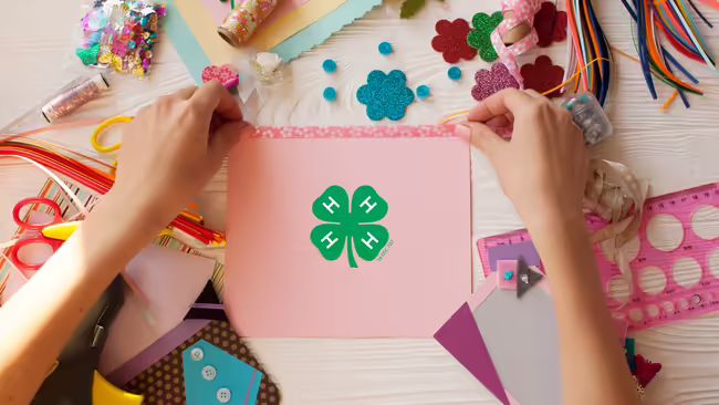 Hands arranging tape on pink paper featuring a green 4-H emblem, surrounded by scrapbooking supplies like ribbon, colorful paper, sequins, and scissors.