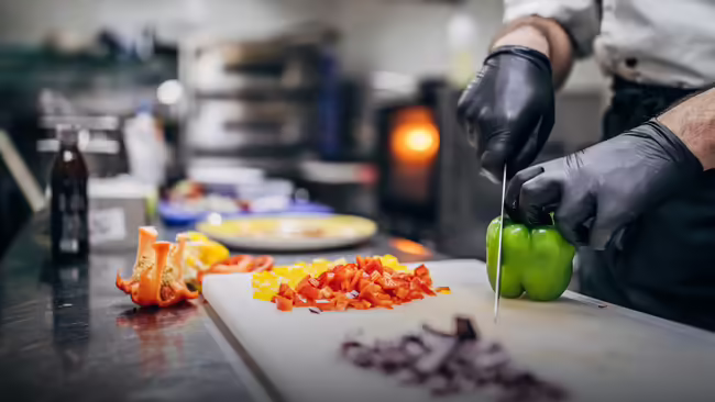 A chef cutting a green pepper on a cutting board with a variety of vegetables that are cut surrounding the cutting board.