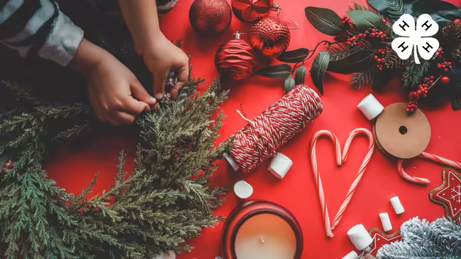 Hands assembling a holiday wreath alongside red ornaments, candy canes, and craft supplies on a red background with a 4-H logo.