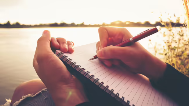 A person writes in a notebook by a lake.