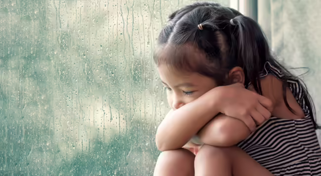Sad preschooler sitting next to a window on a rainy day
