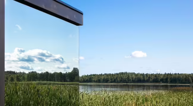 A window reflecting a sky, clouds, a lake, trees, and cattails in its surroundings. 