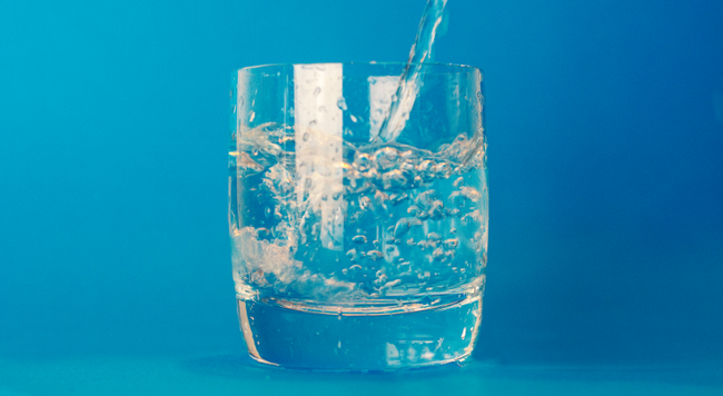 Blue background, glass in the middle of picture with water being poured into it. 