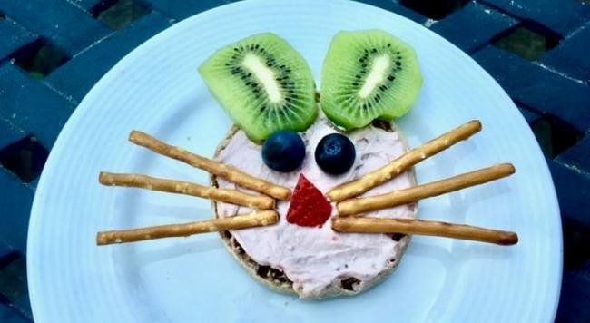 Various pieces of food creating a bunny face (pretzel sticks for whiskers, kiwi slices for ears) on a white plate.