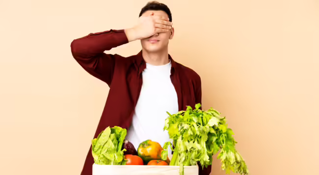Person standing by a crate of vegetables with their hands over their eyes