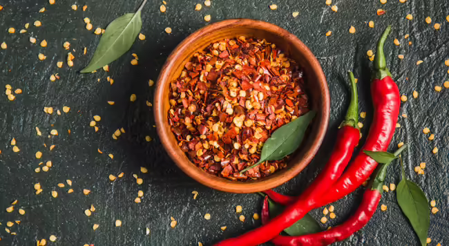 Bowl of red pepper flakes next to 3 red chilis on dark gray background