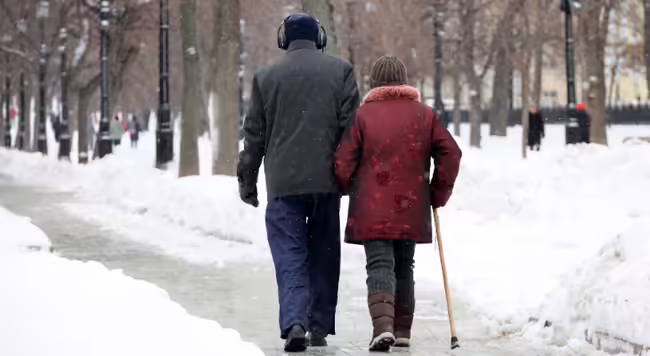An older couple walking outside on a snowy sidewalk. One of them is using a walking cane.