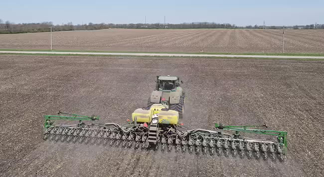 A large corn planter works in a field with blue sky and a road visible in the background.