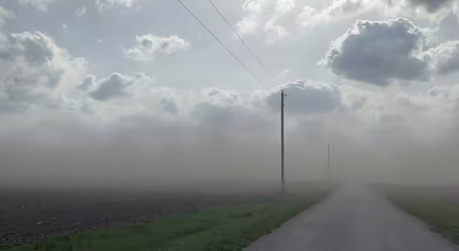 Sunshine peeks through large clouds over thick dust, which shrouds a country road running between two farm fields.