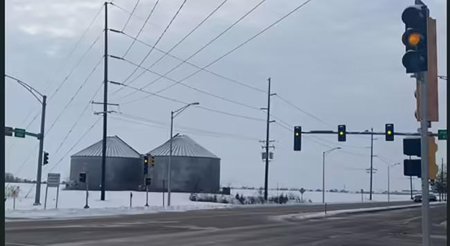 Two large grain bins on snow covered ground are visible at a stoplight intersection with a yellow light.