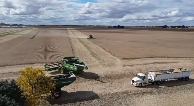 A large farming harvest operating is taking place in an agricultural field with clouds & blue sky in background.