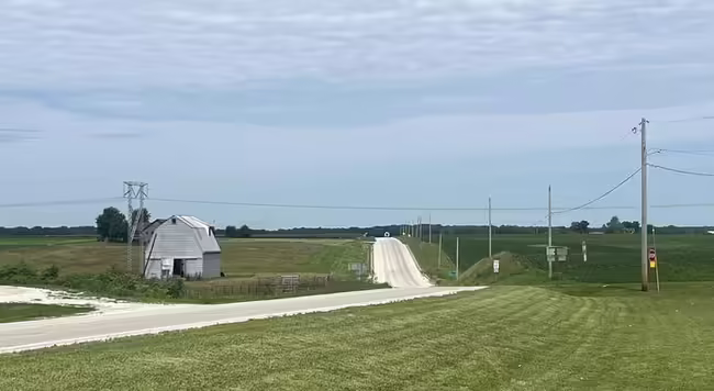 Distant view of a country road with a barn on the left, power lines on the right and a blue sky with some clouds in the background.