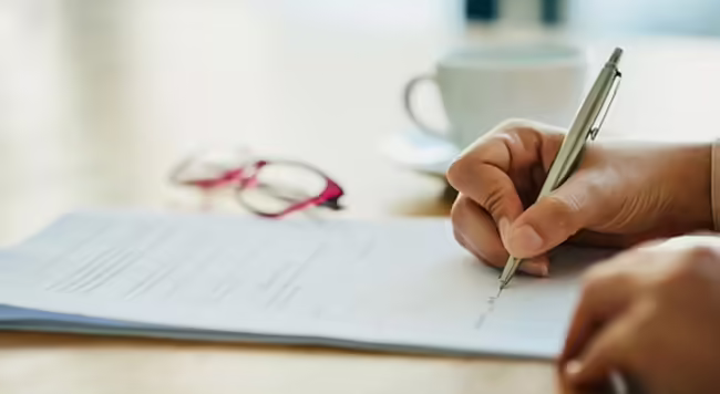 A person's right hand holds a pen to a pad of paper, with glasses and mug off to side.