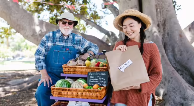 A woman in knitted sweater holding paper bag with a famer both standing next to woven basket filled with fruits