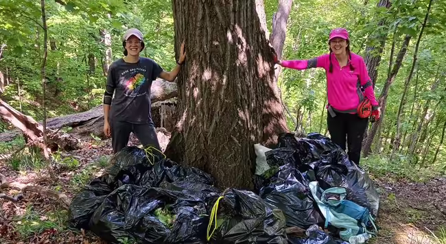 two ladies standing in the woods with five garbage bags full of garlic mustard