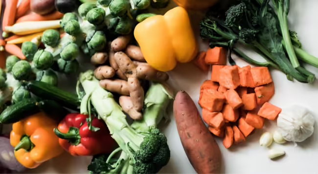 A spread of different vegetables including brussel sprouts, carrots, bell peppers, potatoes, and garlic on a white table.
