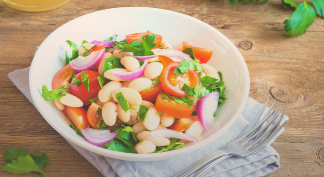 Image of a bowl with tomatoes, red onions, and white beans, next a fork and napkin on a wooden tabletop