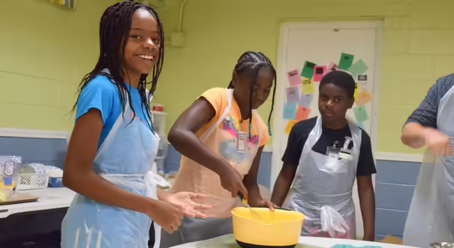 children with aprons, one is stirring food in a bowl.