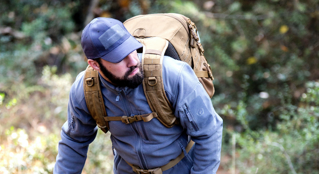 White man with dark beard in blue shirt and hat hiking with backpack