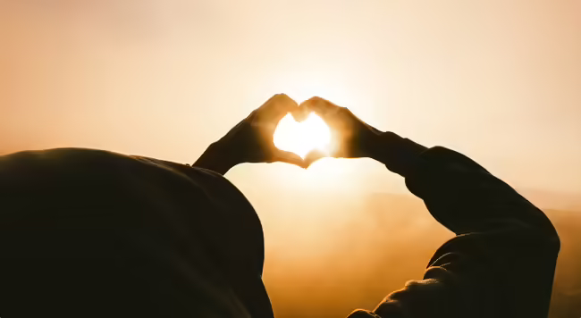 silhouette of person in hooded sweatshirt facing the sun with hands forming a heart