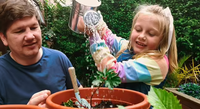 A child watering a plant with an adult watching 