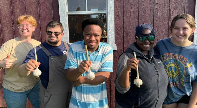 Two Rooted for Good employees stand next to 3 people proudly showing off their garlic harvest