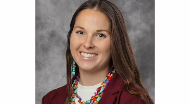 close up of a person smiling with long brown hair, colorful necklace, and maroon jacket