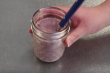 Small mason jar with smoothie and metal spoon with blue handle. Jar is held by a Caucasian hand