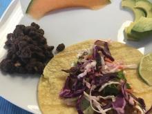 Fish taco on plate with cantaloupe, black beans and avocado slices