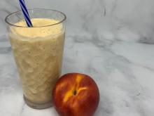 Tall clear glass with smoothie and a straw with a whole peach sitting at base of glass