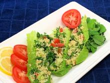 Tabbouleh salad sits inside lettuce leaves on a rectangular white platter. Platter is decorated with slices of lemon and tomato. Background is a blue placemat.